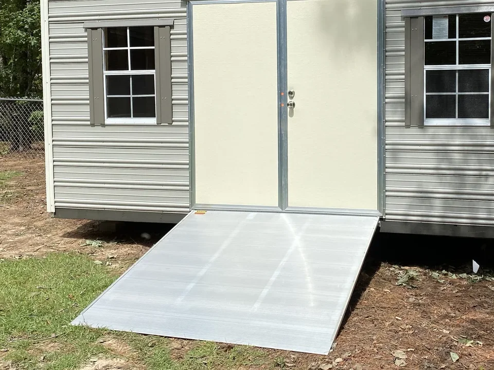 Clean aluminum shed ramp attached at the double doors of a gray shed, showing the full non-slip ramp surface